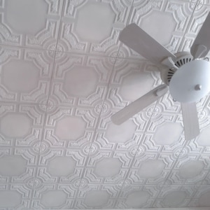May include: White ceiling fan with a white ceiling with a decorative pattern. The fan has five blades and a white light fixture.