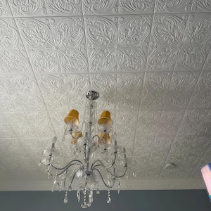 May include: A crystal chandelier with yellow lampshades hangs from a white ceiling with an embossed pattern.