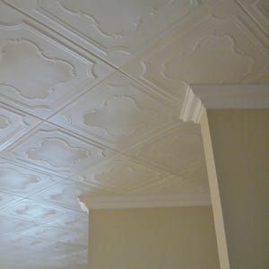 May include: A close-up view of a white, ornate ceiling with a repeating pattern of square tiles. Each tile features a raised, decorative design with a fan-like motif. The ceiling is adjacent to a white crown molding and a light beige wall.