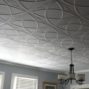 May include: White, textured ceiling tiles with a repeating geometric pattern. The design features intersecting circles and diamond shapes. A chandelier hangs from the ceiling, and a window with blinds is visible in the background.