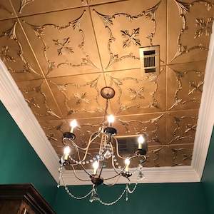 May include: A gold-colored ceiling with ornate, embossed floral patterns. A decorative chandelier with crystal accents hangs below. The ceiling is framed by a white molding, and the wall is painted a deep teal.