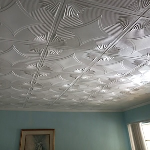 May include: A white, textured ceiling with a repeating pattern of stylized sunbursts and curved lines. The ceiling tiles are arranged in a grid, creating a decorative and architectural element. The room's walls are painted a light blue.