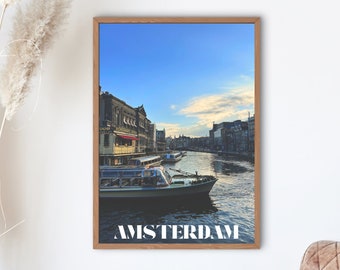 Könnte beinhalten: Ein gerahmtes Foto eines Kanals in Amsterdam, Niederlande. Das Foto zeigt einen Kanal mit Booten und Gebäuden auf beiden Seiten. Der Text "AMSTERDAM" ist in Weiß am unteren Rand des Fotos gedruckt.