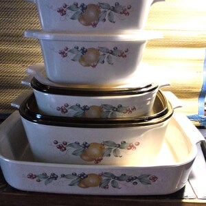 May include: A stack of five off-white rectangular ceramic baking dishes with integrated side handles. Each dish features a central botanical motif of a yellow pear surrounded by green leaves, dark blue berries, and small red berries on the front side.