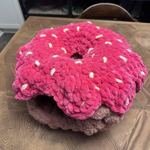 May include: A round, plush, crocheted doughnut-shaped pet bed featuring a thick, textured dark pink top layer with scattered white yarn sprinkles and a solid brown base layer. The design mimics a frosted doughnut with a central opening in the middle.