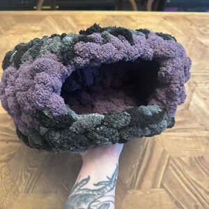 May include: A round, chunky crochet pet bed or cave featuring a variegated yarn pattern in shades of deep purple, charcoal gray, and black. The thick, textured yarn creates a soft, rounded structure with a wide, open entrance for a small animal.