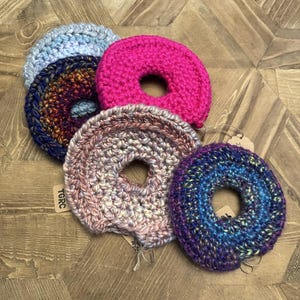 Könnte beinhalten: Fünf kreisförmige, handgehäkelte Haargummis in verschiedenen Farben und Texturen. Die Kollektion umfasst ein einfarbiges leuchtendes Pink, ein Hellblau, eine mehrfarbige Mischung aus Dunkelblau und Gold, Beige und Grau sowie Blau und Violett.