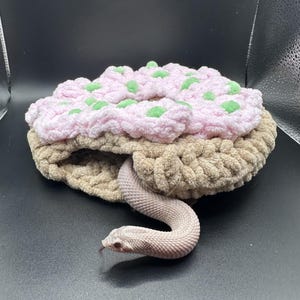 Op de afbeelding: Een lichtroze en beige gehaakt accessoire in de vorm van een donut met kleine groene accenten aan de bovenkant. Een bleke slang met gladde schubben en een zichtbare tong komt uit de centrale opening van het gehaakte object naar buiten.