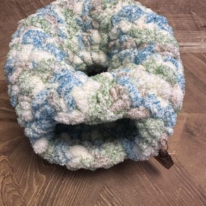 May include: A round, thick, chunky knit cowl scarf featuring a variegated yarn pattern in shades of white, light blue, sage green, and soft beige. The textured, plush accessory has a circular opening and a small brown tag attached to the edge.