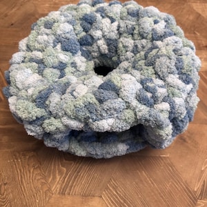 Puede incluir: Un cojín circular con forma de rosquilla hecho de hilo grueso y voluminoso. El tejido presenta una paleta de colores variados que consiste en varios tonos de azul, verde salvia y blanco, creando una apariencia suave, texturizada y tonal.
