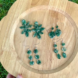 Puede incluir: Tres pares de pendientes colgantes con patrones de mármol verde. Un par tiene componentes en forma de hoja, un par presenta dijes en forma de diamante con pequeños tachuelas verdes y un par consta de tres cuentas circulares apiladas.