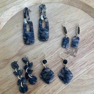 May include: A collection of handmade earrings in various designs. The earrings feature a marbled pattern in shades of gray and blue. Styles include rectangular, geometric, and chain link designs. Some earrings have gold-colored hooks and accents.