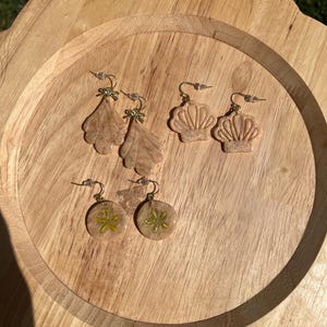 May include: A collection of handmade earrings in various designs. The earrings include leaf, seashell, and flower shapes, all in neutral tones with gold-colored accents. The earrings are displayed on a wooden tray.