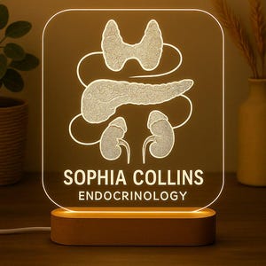 Puede incluir: Una luz de noche personalizada con una base de madera. El panel acrílico transparente presenta una ilustración blanca brillante de órganos humanos, incluyendo la tiroides, el páncreas y los riñones. El nombre "SOPHIA COLLINS" y la palabra "ENDOCRINOLOGY" están impresos debajo.