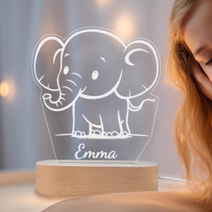 以下が含まれることがあります： 漫画の象のデザインと「Emma」という名前が下に印刷された白いアクリル製のナイトライト。ナイトライトは木製のベースの上に置かれています。