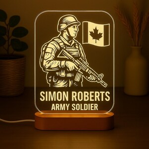 May include: A personalized night light featuring a soldier illustration with a Canadian flag. The light has a wooden base and a clear acrylic panel with the name "SIMON ROBERTS" and the text "ARMY SOLDIER" engraved on it.