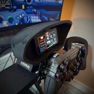 May include: A black racing simulator setup featuring a steering wheel and a display screen. The screen shows racing data with the text "MOZA RACING" visible. The steering wheel has multiple buttons and knobs, and the setup is mounted on a black frame.