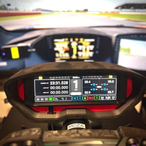以下が含まれることがあります： レースシミュレーターのダッシュボードのクローズアップ。ラップタイムやその他のパフォーマンス指標を表示するデジタルディスプレイがあります。ダッシュボードは黒いスエードのようなフードとステアリングホイールを備えています。「MOZA」のブランド名が見えます。