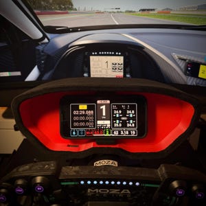 May include: A close-up of a racing simulator dashboard. The dashboard features a digital display with various data points, including lap times and speed. The display is set in a red and black frame, with the MOZA Racing logo visible.