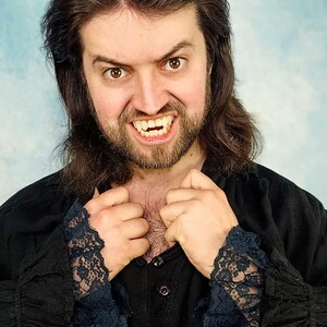 May include: A person with long brown hair and a beard, wearing a black shirt with lace cuffs. The person is baring their teeth, with visible fangs. The background is a light blue color.
