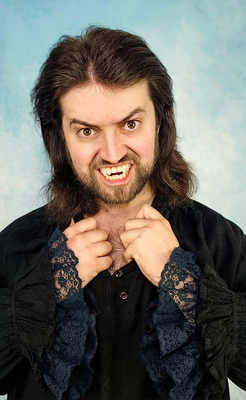 May include: A person with long brown hair and a beard, wearing a black shirt with lace cuffs. The person is baring their teeth, with visible fangs. The background is a light blue color.