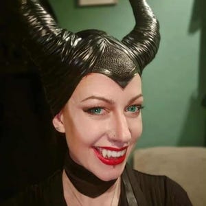 May include: A person wearing a black, textured, Maleficent-style headpiece with large, curved horns. The person has green eyes, red lipstick, and vampire fangs. A black choker and necklace are also visible.
