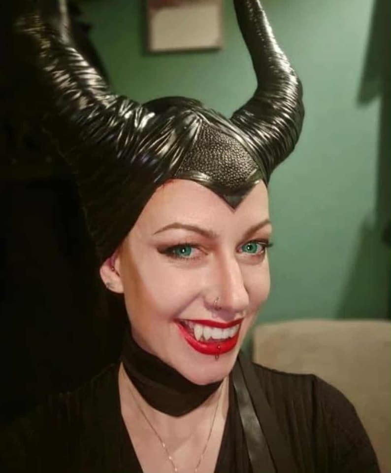 May include: A person wearing a black, textured, Maleficent-style headpiece with large, curved horns. The person has green eyes, red lipstick, and vampire fangs. A black choker and necklace are also visible.