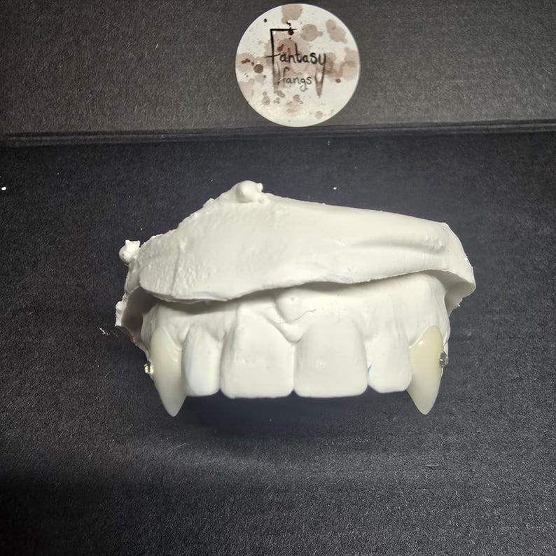 May include: White, custom-made vampire fangs. The fangs are made of a white material and are designed to fit over the natural teeth. The image also includes a round sticker with the text "Fantasy Fangs".