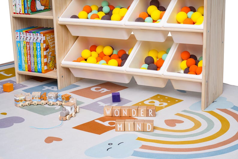 May include: A wooden toy storage unit with white bins filled with colorful balls. A wooden train and blocks spelling "WONDER MIND" sit on a colorful play mat with a rainbow design. Bookshelves are visible.