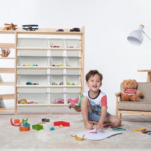 May include: A wooden toy storage unit with multiple shelves and white bins, filled with toys. A young child is drawing on the floor with crayons, surrounded by toys. A teddy bear sits in a small chair.