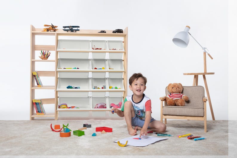 May include: A wooden toy storage unit with multiple shelves and white bins, filled with toys. A young child is drawing on the floor with crayons, surrounded by toys. A teddy bear sits in a small chair.