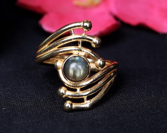 Può includere: Anello dorato con una pietra centrale rotonda e iridescente. L'anello presenta un design aperto con bande curve e parallele e piccoli accenti sferici. L'anello è posto su uno sfondo nero con un fiore rosa sfocato.