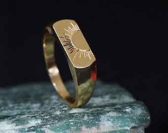 May include: A gold-colored ring with a rectangular face featuring a sunburst design. The ring is set against a dark background, highlighting its polished finish and intricate details. The ring's band is smooth and rounded.
