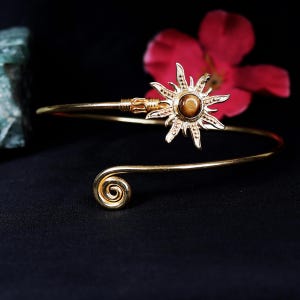May include: Gold arm cuff with a sunburst design and a central brown stone. The cuff has a spiral detail at one end and a decorative element near the sunburst. The background is black with a red flower.