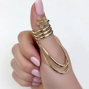 May include: A gold-colored finger ring set. The ring features a floral design on the top finger and multiple bands wrapping around the finger. Two curved gold bands extend down the side of the finger. The ring is worn on a hand with pink nail polish.