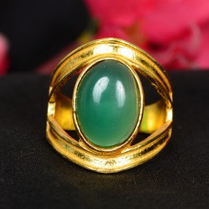 May include: A gold ring with a large, oval-shaped green gemstone set in a gold bezel. The ring has a wide band with a decorative design.
