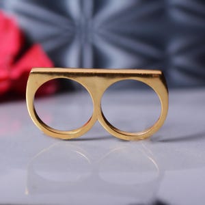 Puede incluir: Un anillo de doble dedo de oro con una banda ancha. El anillo tiene dos círculos conectados.