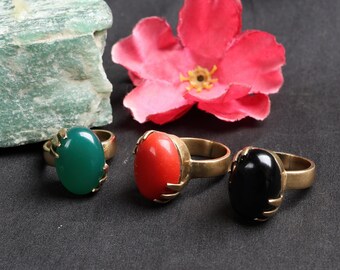 May include: Three gemstone rings with oval-shaped stones in green, red, and black. The rings have a gold-colored band and setting. A pink flower and a piece of green stone are in the background.
