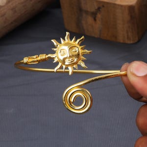 May include: A gold-toned metal cuff bracelet featuring a central sun motif with a human face and radiating rays. One end of the open band terminates in the sun design, while the other end features a simple, circular spiral coil detail.