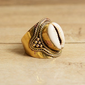 May include: A gold ring featuring a white seashell design. The ring has an intricate, ornate design with a rope-like pattern and small gold beads.