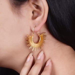 Può includere: Orecchini a cerchio dorati con un design a forma di sole. Gli orecchini sono in metallo e hanno una finitura lucida.