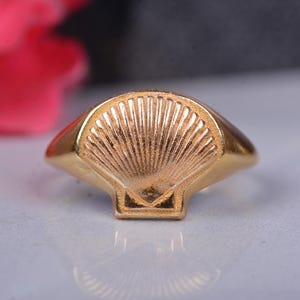Può includere: Un anello d'oro con un designo a conchiglia dettagliato. L'anello è una fascia semplice con un designo a conchiglia in rilievo al centro.