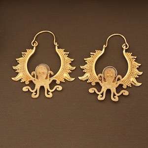 May include: A pair of gold-colored octopus earrings. Each earring features an octopus design in the center, surrounded by a sunburst-like frame. The earrings have a hook closure and are set against a brown background.