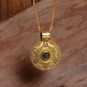 Op de afbeelding: Een goudkleurige hangerketting met een cirkelvormig ontwerp. De hanger heeft een centrale steen en ingewikkelde details rond de randen. De ketting is ook goudkleurig, wat een samenhangende uitstraling creëert. De ketting wordt gepresenteerd tegen een houten achtergrond.