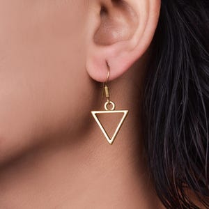 Peut inclure: Boucles d'oreilles en forme de triangle dorées avec un design géométrique simple. Les boucles d'oreilles pendent à l'oreille d'une personne.