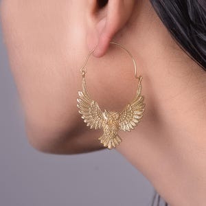 Op de afbeelding: Gouden oorringen met een uil-ontwerp. De uil heeft gedetailleerde vleugels en een getextureerd lichaam. De oorbellen hangen aan een cirkelvormige ring. Het ontwerp is ingewikkeld en elegant, geschikt om een vleugje natuurlijke stijl toe te voegen.