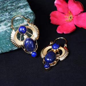 Könnte beinhalten: Ein Paar goldfarbene Ohrringe mit einem einzigartigen Design. Jeder Ohrring hat drei blaue Edelsteine: einen kleinen runden oben, einen ovalen in der Mitte und einen kleinen runden unten. Die Ohrringe haben ein dekoratives, durchbrochenes Design.