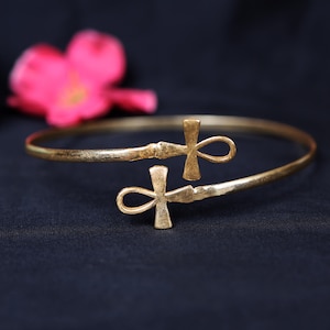May include: A gold-coloured cuff bracelet featuring two ankh symbols. The bracelet rests on a dark blue surface, with a blurred pink flower in the background. The ankh symbols are a key element of the bracelet's design.