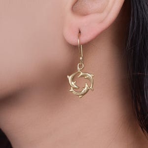 Könnte beinhalten: Goldfarbene Ohrringe mit einem Design aus drei Delfinen, die im Kreis schwimmen.