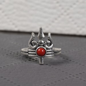 Op de afbeelding: Een zilverkleurige metalen ring met een drietandvormig middendeel en een kleine, ronde, felrode steen aan de basis. Het metaal heeft een antieke afwerking met zichtbare details op de ringband en het ontwerp van de drietand met drie punten.